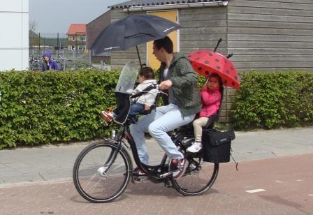 Man cycling in the rain with two young children on his bike, both under umbrellas, on a Dutch cycle path