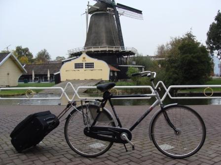 Bicycle with a wheeled suitcase strapped on, parked in front of a Dutch windmill