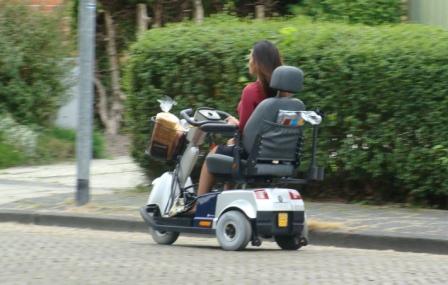 Person riding a three-wheeled mobility scooter along a street beside a hedge