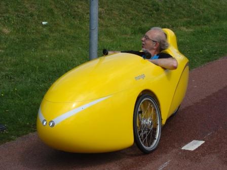 Man riding a streamlined yellow recumbent velomobile along a cycle track