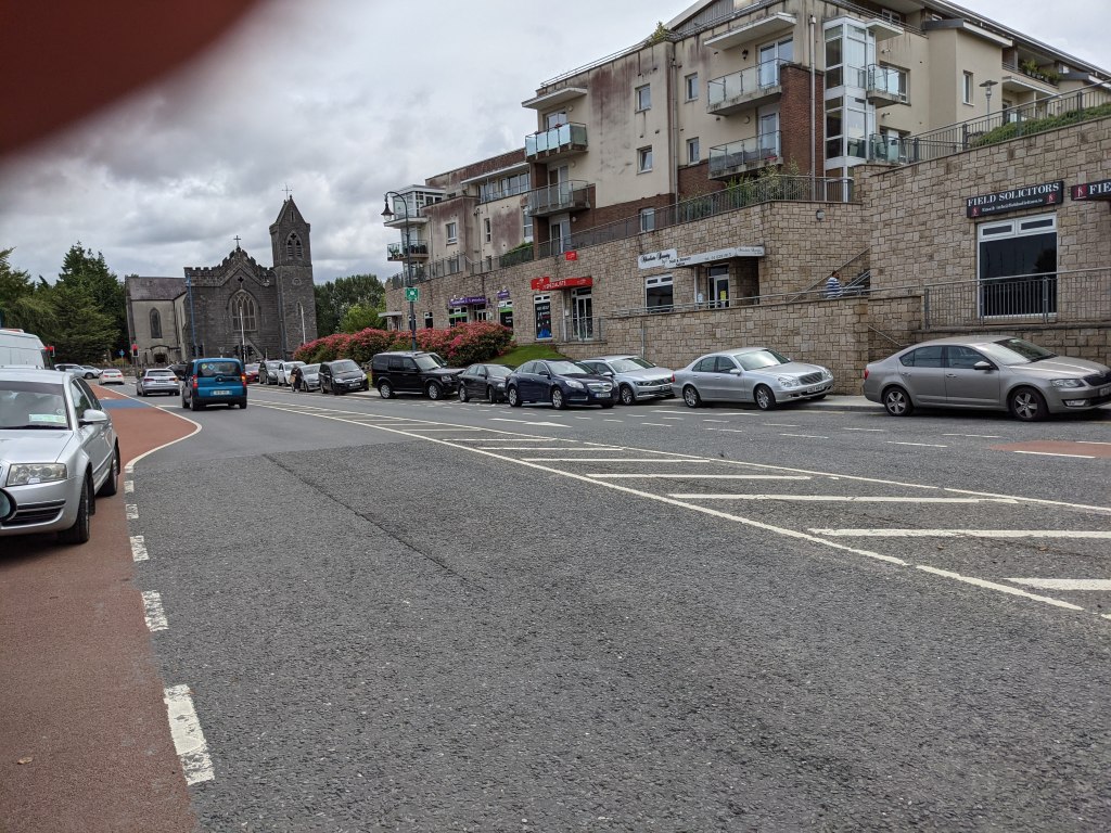 A long row of cars parked along the shared path on the Kilcock Road, with the church and apartments behind
