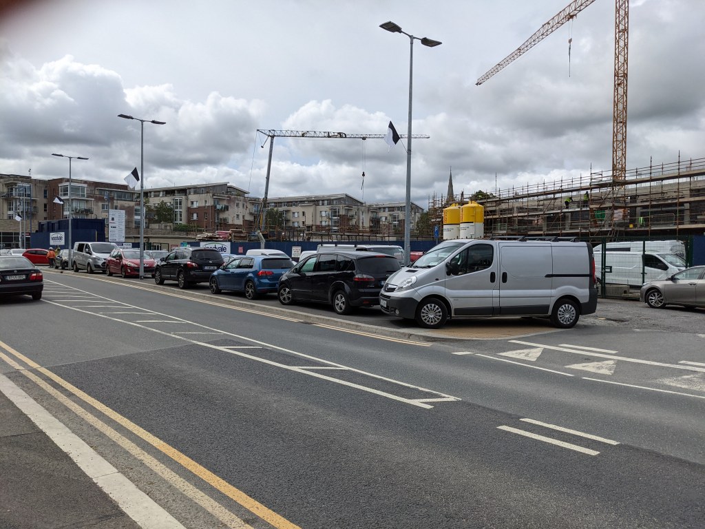 Cars and vans parked along the Moyglare Road beside a construction site with cranes