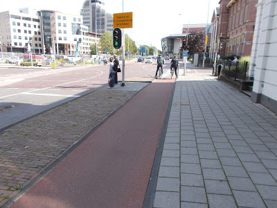 Red asphalt cycle track beside a grey paved footpath at a Dutch urban junction, with cyclists and pedestrians