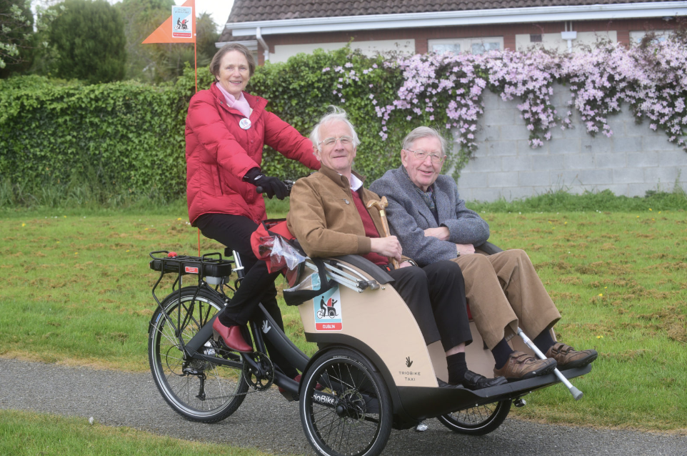 A volunteer pilots a Cycling Without Age trishaw carrying two elderly passengers on its front bench