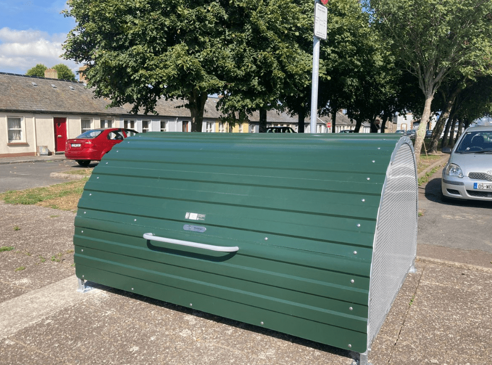 A green lockable on-street bike hanger with a curved roof, sited in a former car parking space