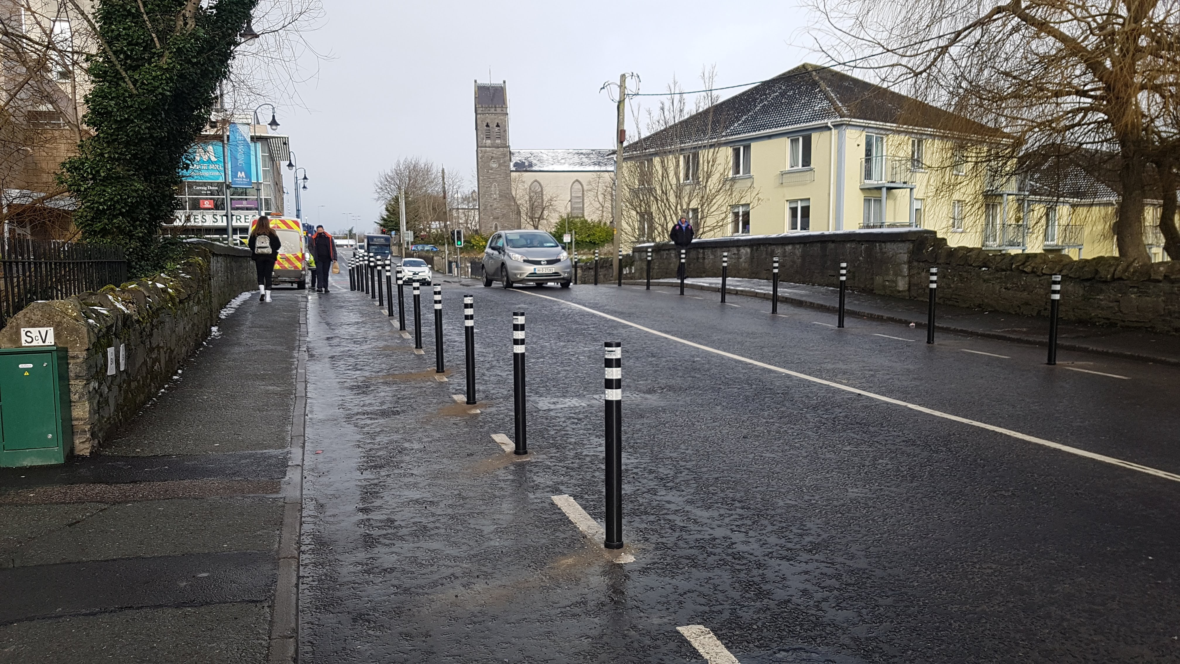 Black-and-white bollards separate a new pop-up cycle lane from traffic on a wet Maynooth street, church tower behind.