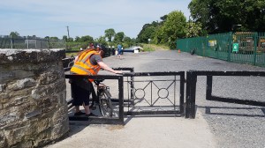 Person in a hi-vis vest pushing a bicycle through a metal kissing gate on a greenway, sports pitch behind