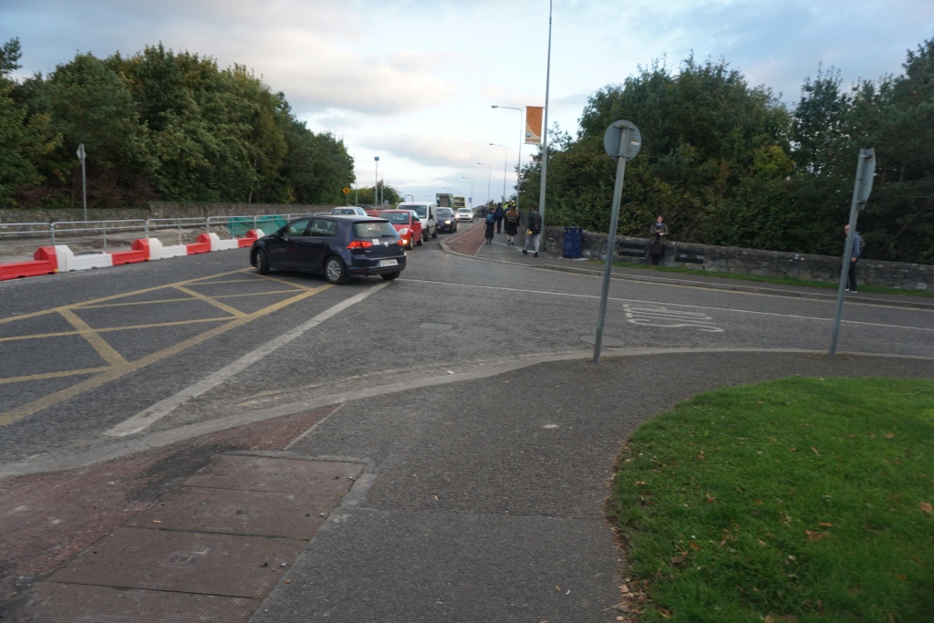Cars queue past red-and-white barriers narrowing Maynooth’s Straffan Road during roadworks, pedestrians on the footpath