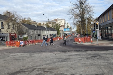 Pedestrians cross a torn-up Mill Street, Maynooth, amid roadworks lined with orange barriers and town-centre buildings.