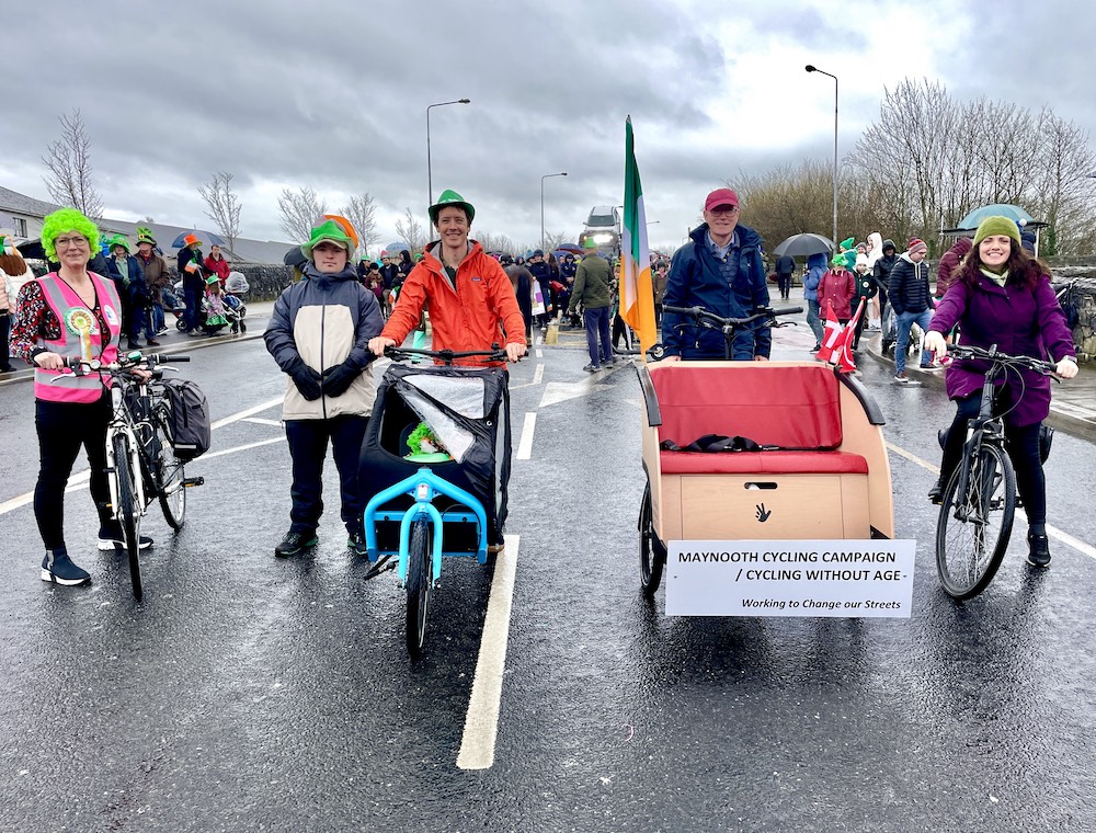 Six MCC members in green St. Patrick’s hats pose across the road with bikes, a cargo bike and the trishaw