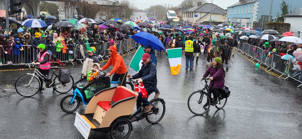 Cyclists and the Cycling Without Age trishaw in Maynooth’s rainy St. Patrick’s Day parade, crowds with umbrellas