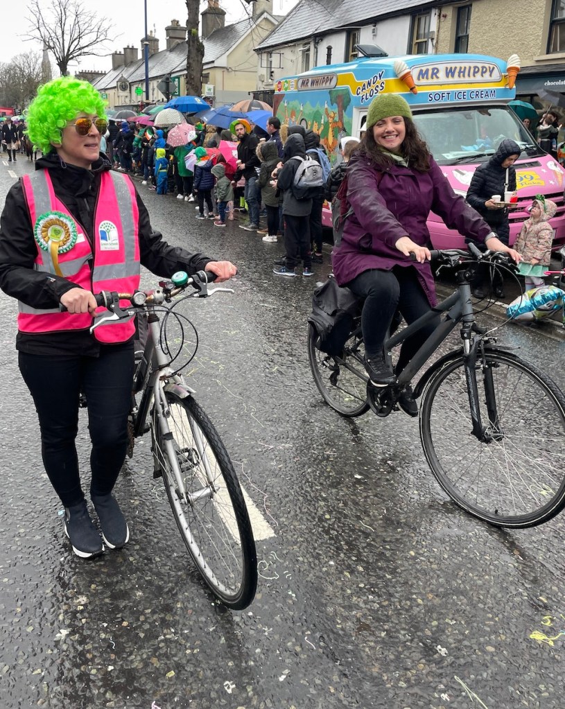 Two women cycle the parade route past a Mr Whippy ice cream van, one in a green wig and pink hi-vis vest