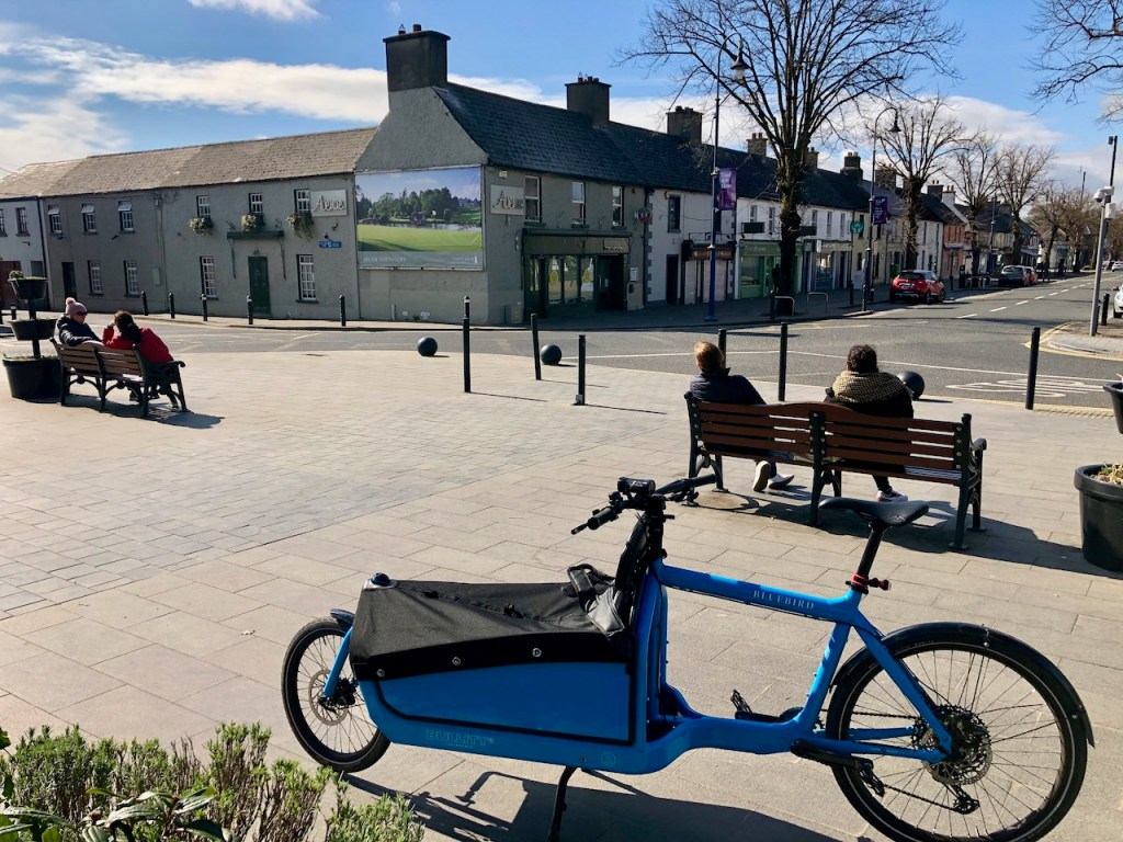 Blue cargo bike parked at the top of Maynooth’s Main Street, with people resting on benches and town shops behind