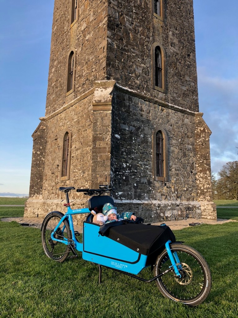 Blue Bullitt cargo bike with a bundled-up toddler in the box, parked on grass before the tall stone Tyrconnell Tower