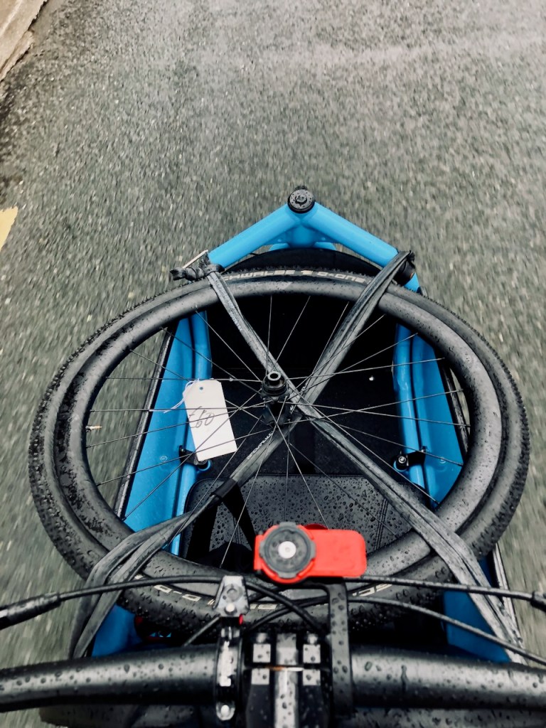 Looking down from the rider’s seat into the cargo box carrying two road-bike wheels on a wet road