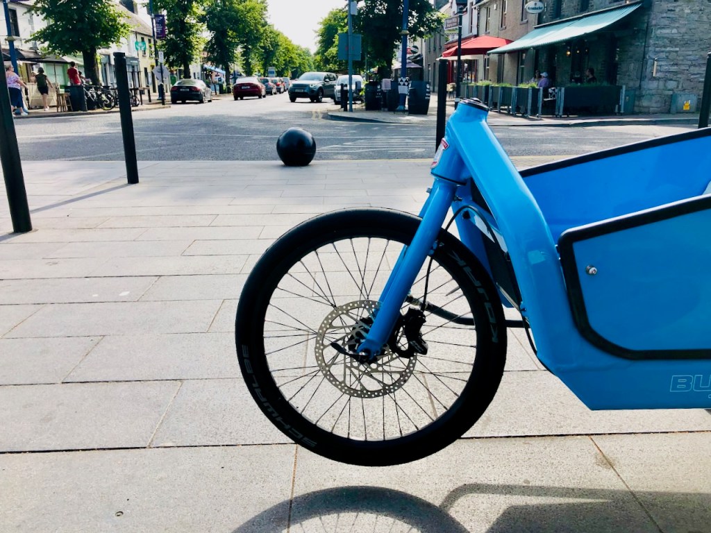 Close-up of the blue cargo bike’s front wheel and disc brake, with Maynooth’s Main Street shops behind