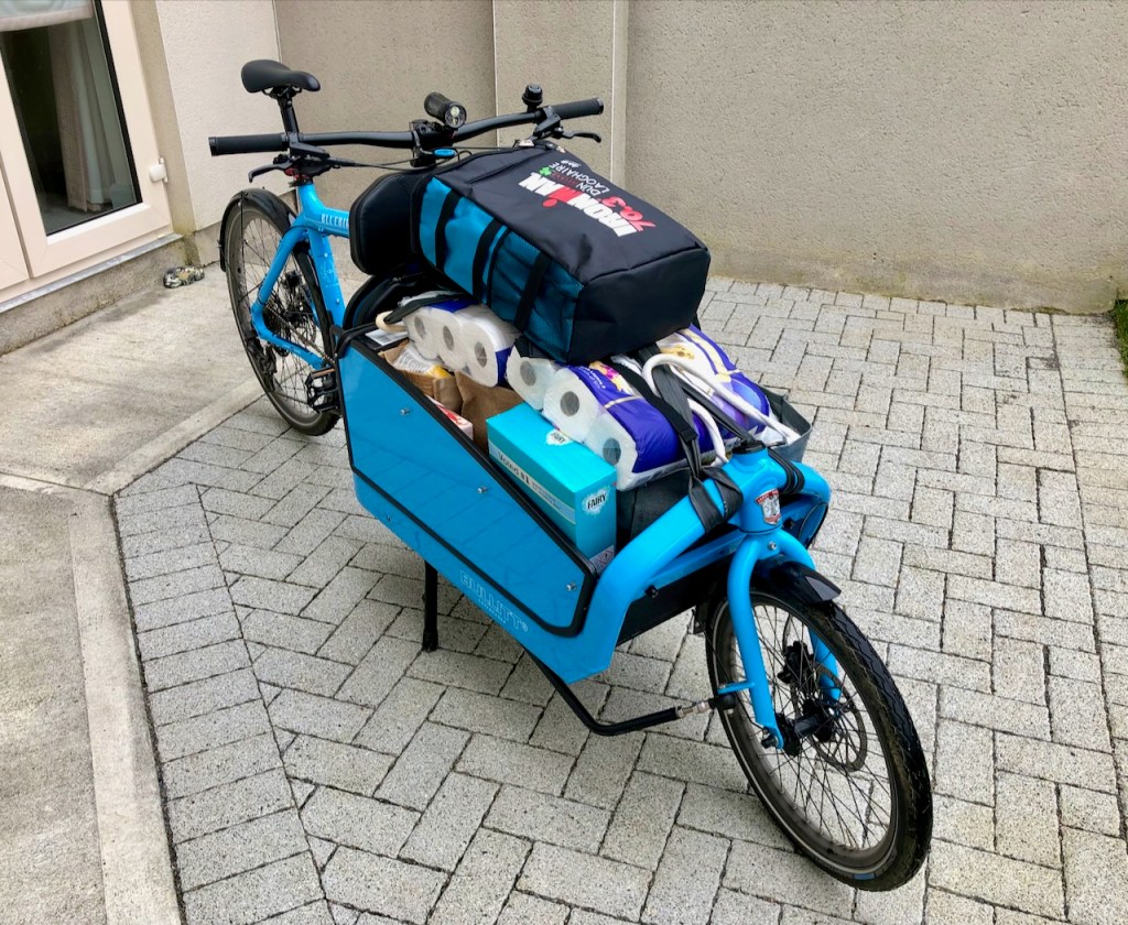 Blue cargo bike with its box full of groceries - toilet roll, tissues and bags - plus an insulated bag behind the saddle