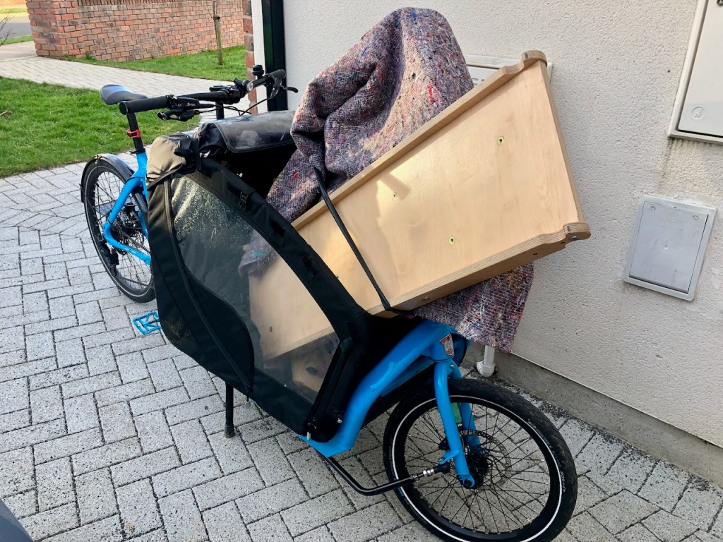 Blue cargo bike loaded with a large wooden cabinet and a moving blanket, parked beside a house