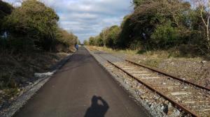 Straight tarmac greenway stretching ahead with rusted disused rails alongside under a blue Westmeath sky.
