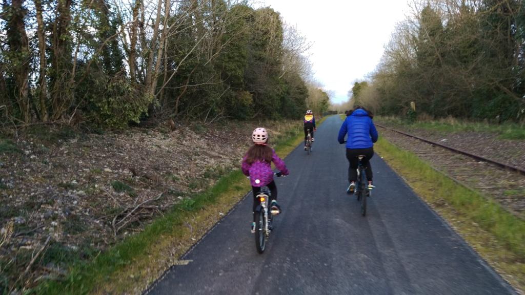 Three cyclists, including a child, ride a smooth tarmac greenway with the disused railway line alongside.