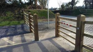 Timber post-and-rail chicane barrier narrowing the greenway entrance where it meets a road.