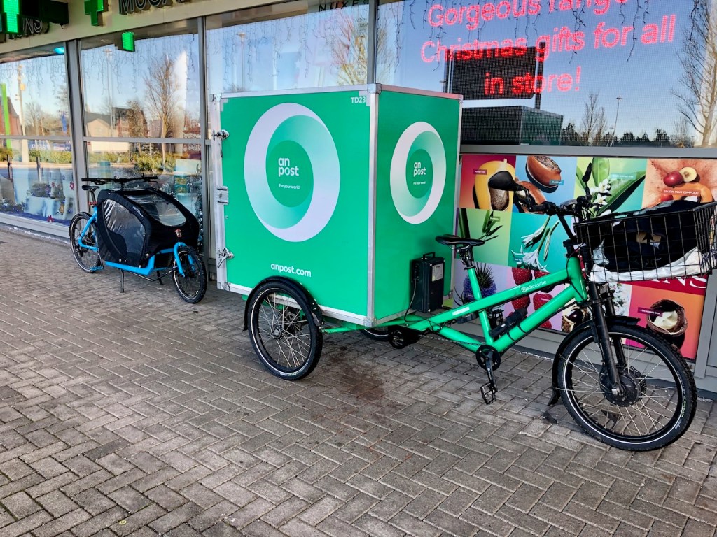 Two cargo bicycles parked beside each other. One is a LvH Bullitt, the other an An Post trike.